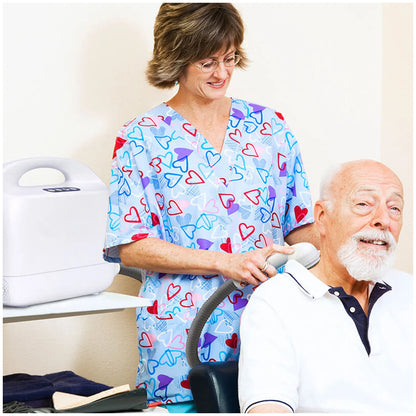 A woman in blue heart-patterned medical scrubs operates a portable shower device on an elderly man seated in a chair in a clinical room.