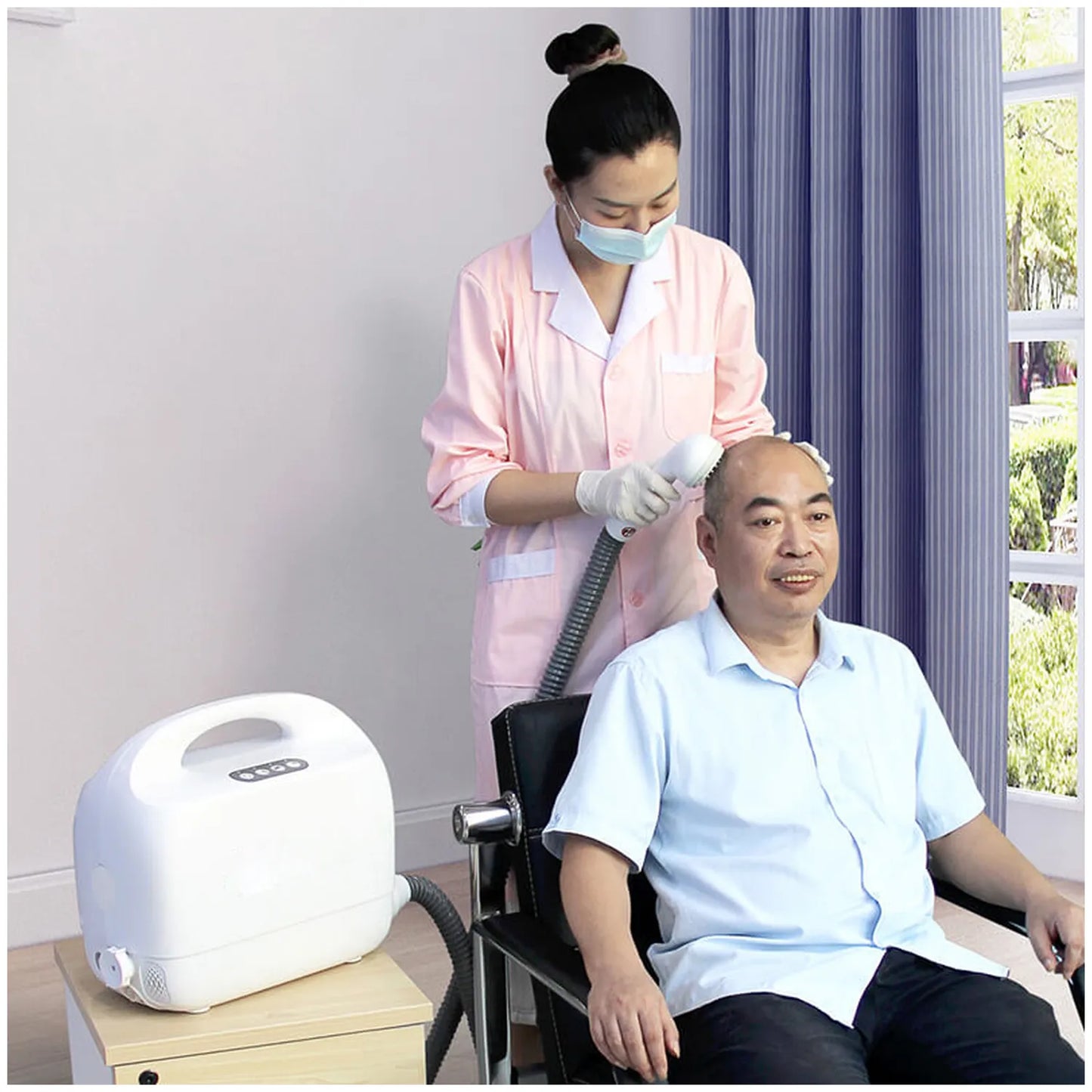 A man sits in a chair while a nurse in a pink uniform uses a handheld portable shower device. The room has light walls, a window with purple curtains, and visible greenery outside.